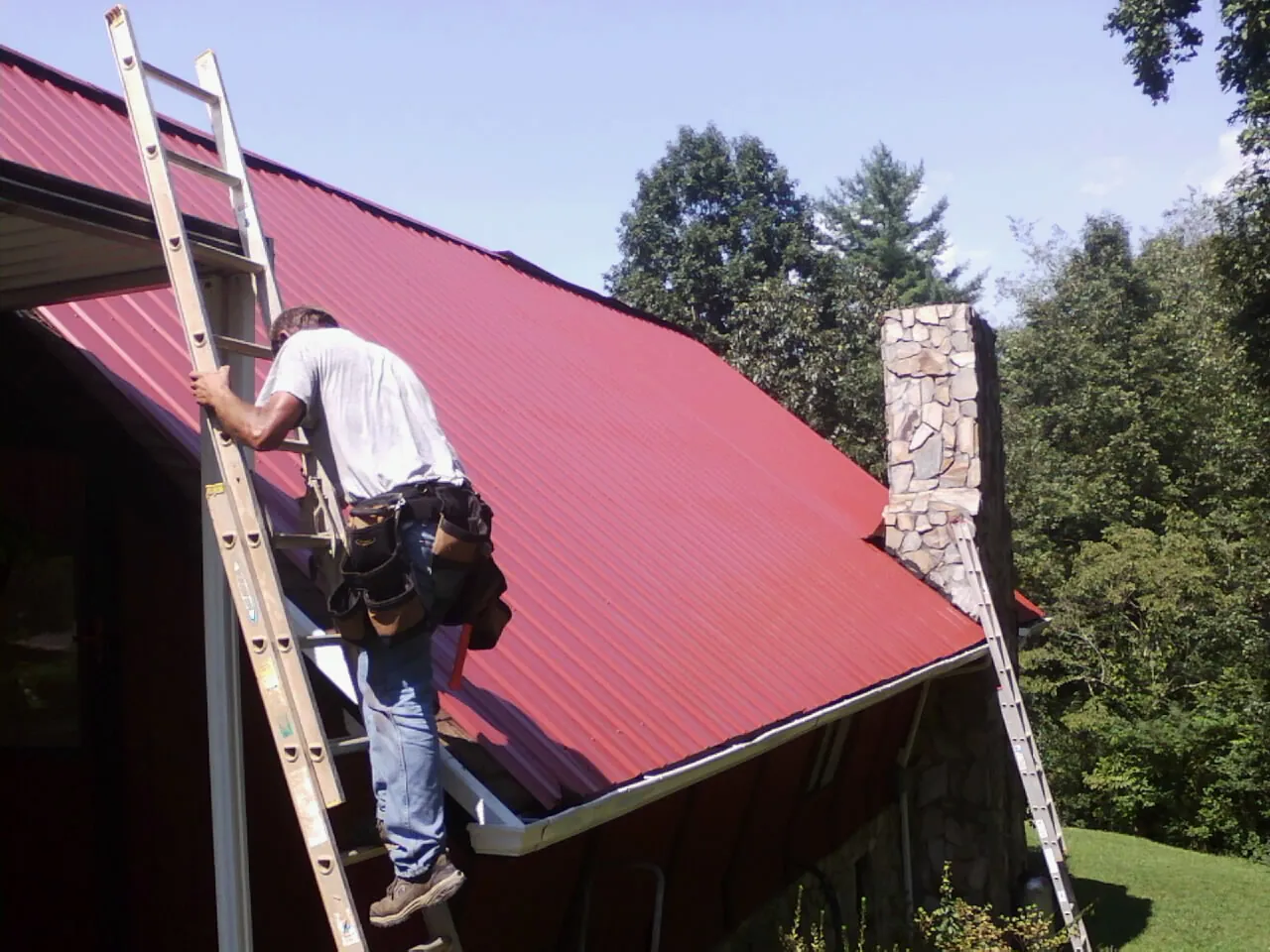 Roofer on a ladder beside a red metal roof and stone chimney — Soffit & Fascia Repair by Roofing Brothers in Rome City